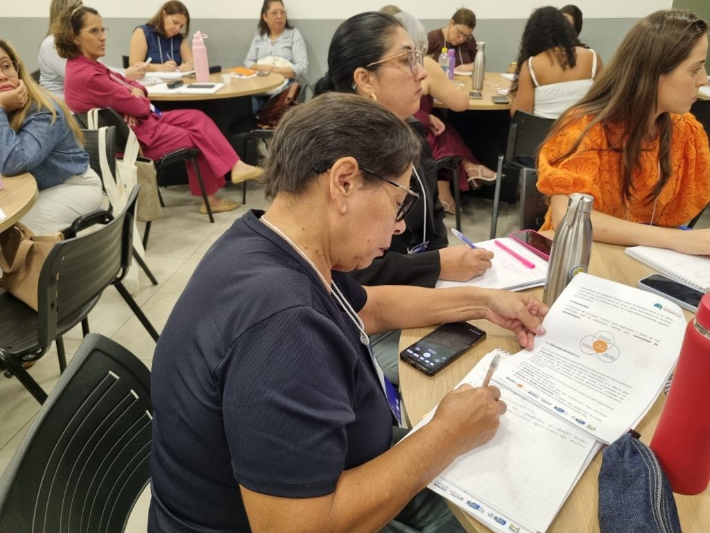 A imagem representa várias pessoas ao redor de várias mesas assistindo os palestrantes falarem sobre as práticas e metodologias inovadoras no ensino de matemática na educação infantil.